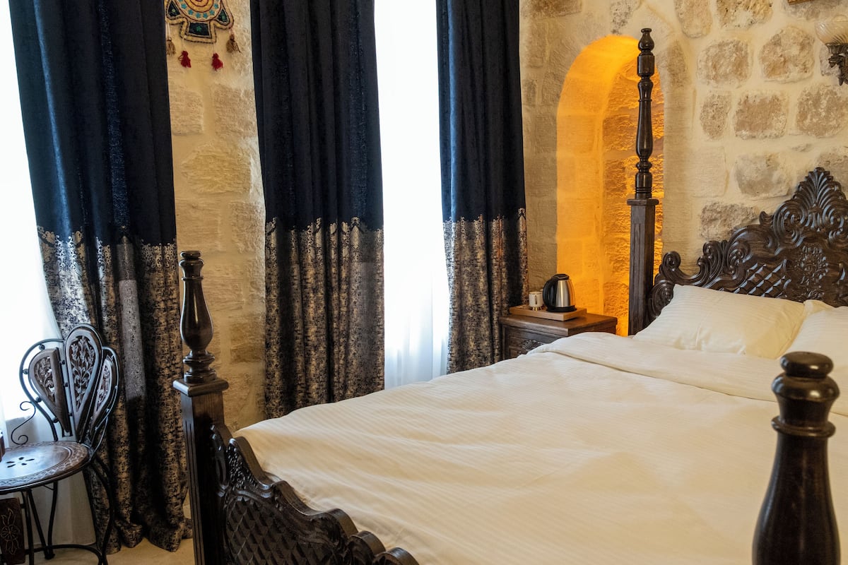 A bedroom with a large bed, dark curtains, a stone wall, and a small table with a chair.