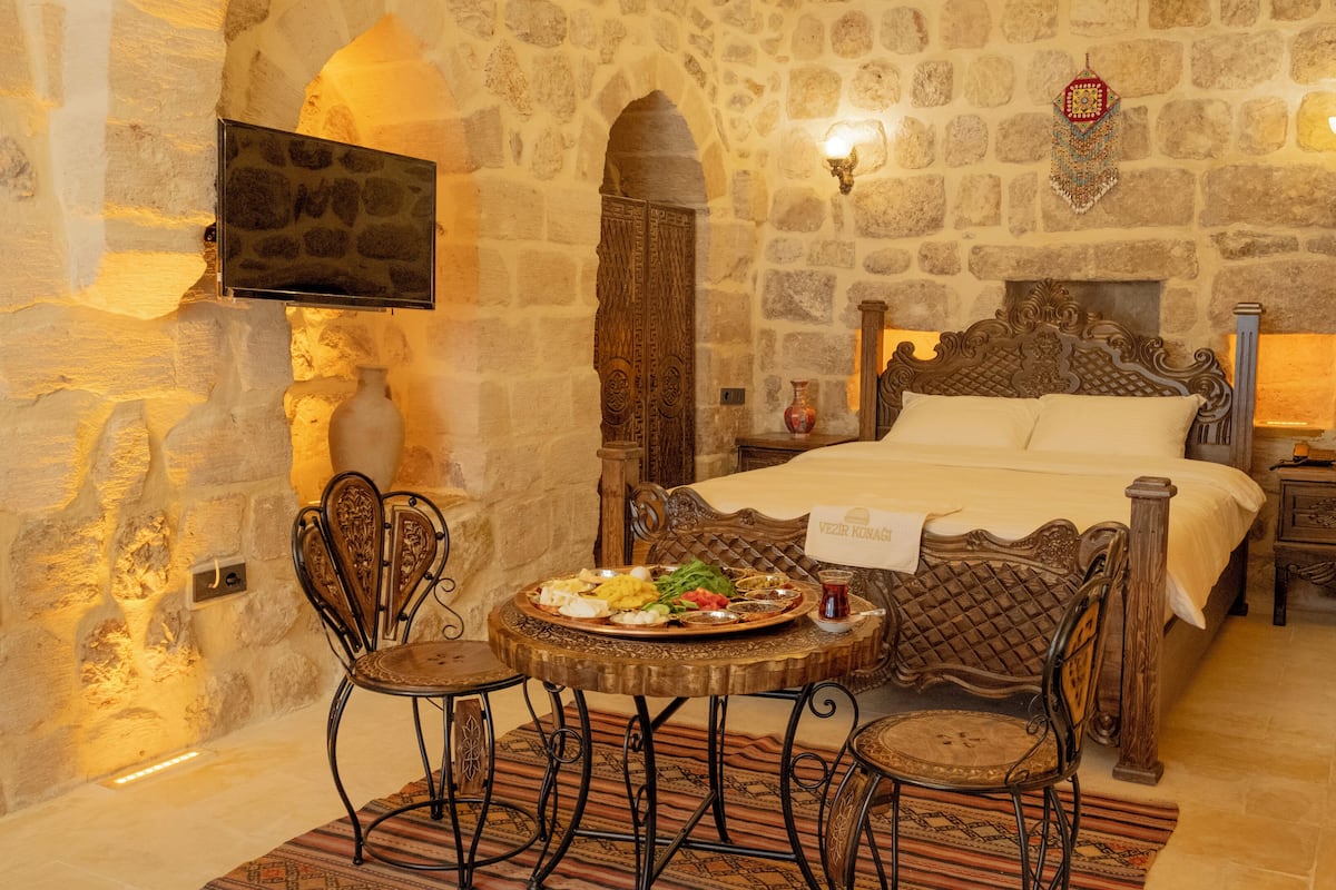 A traditional stone-walled room with a bed, two chairs, a table with food, and a TV mounted on the wall.
