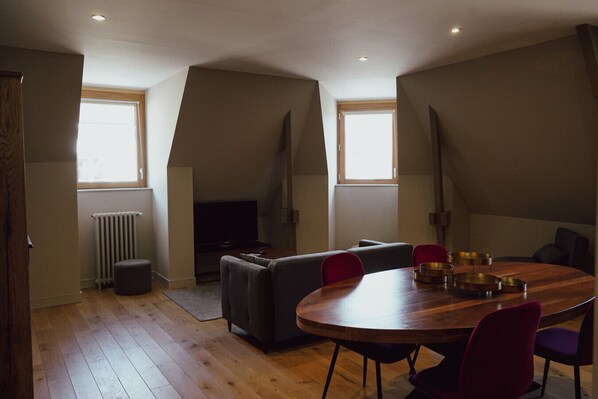 A modern living room with a wooden floor, grey sofa, red chairs, a round dining table with metal bowls, and a TV against the wall.