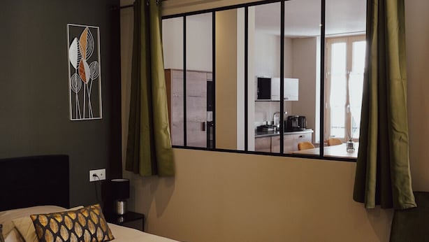 A hotel room with a bed, a chair, a window with curtains, and a view of the kitchen. - Les Résidences Gueguen - Suite Les Clos (Chablis)