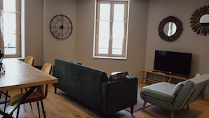 A living room with a wooden dining table, two sofas, a TV, and a large window. - Les Résidences Gueguen - Suite Les Clos (Chablis)
