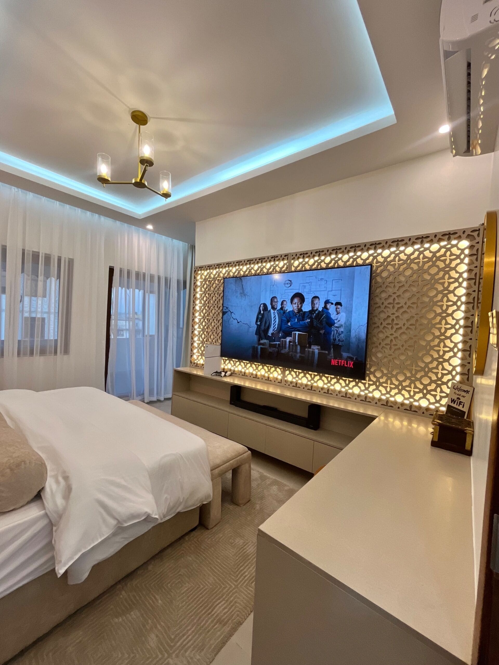 Quarto de hotel moderno com uma televisão de tela grande, fixada em uma parede decorativa, uma cama com lençóis brancos, uma mesa de cabeceira e um ventilador de teto.