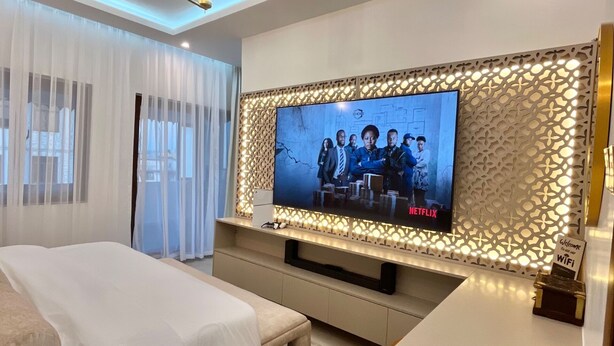 A modern hotel room with a large flat-screen TV mounted on a decorative wall, a bed with white linens, a bedside table, and a ceiling fan. - ZAURA ZANZIBAR (Zanzibar Town)