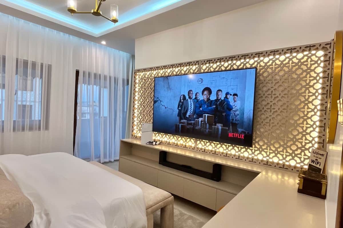 Moderni hotellihuone, jossa on suuri, seinälle asennettu litteänäyttöinen televisio, valkoisilla lakanoiden peitetty sänky, yöpöytä ja kattotuuletin.
