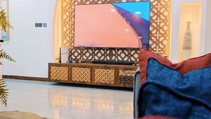 A living room with a flat-screen TV mounted on a decorative wall, a sofa with cushions, a coffee table, and a patterned rug. - ZAURA ZANZIBAR (Zanzibar Town)