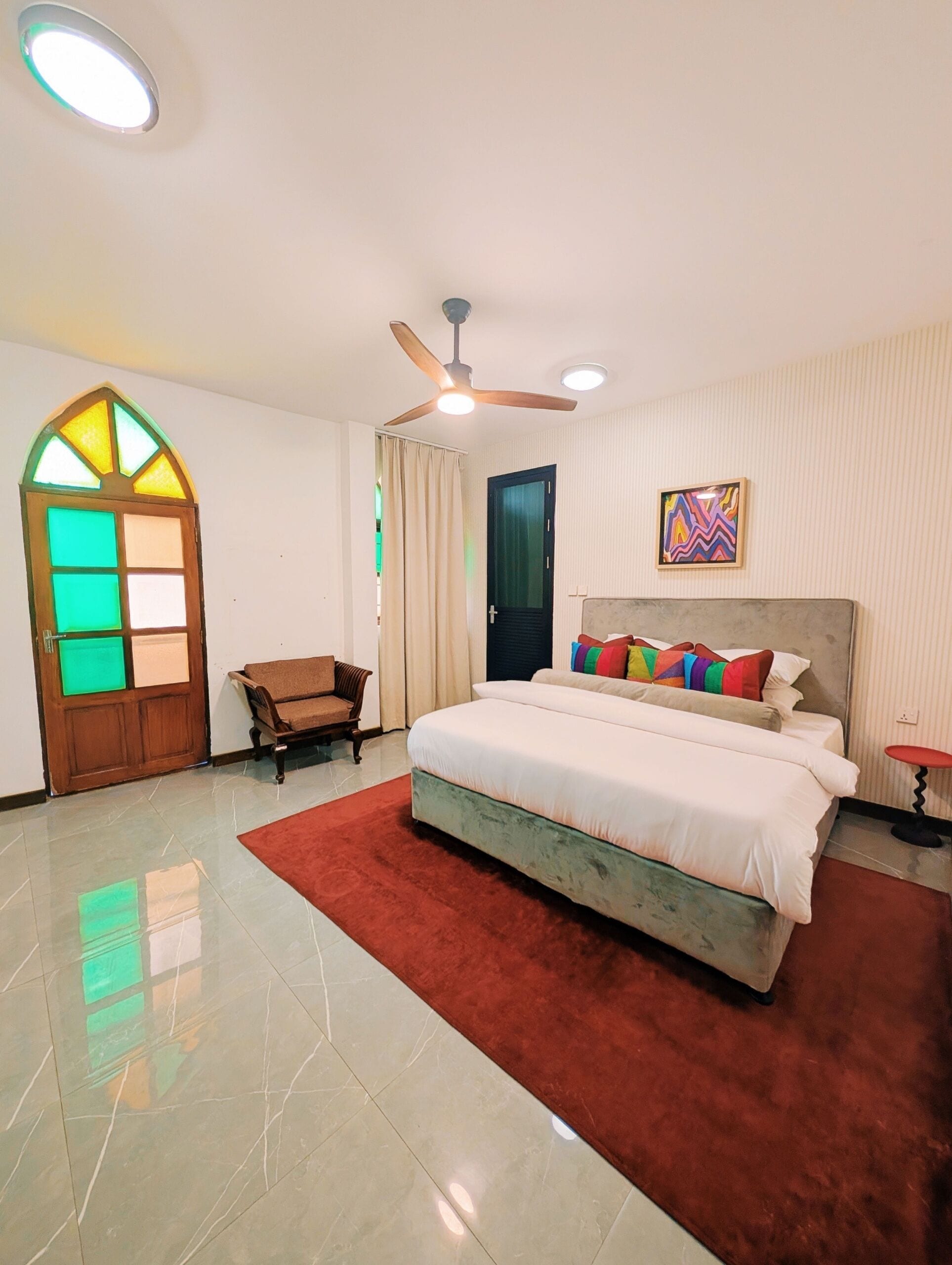 A hotel room with a bed, a ceiling fan, a door with stained glass, a chair, and a painting on the wall.