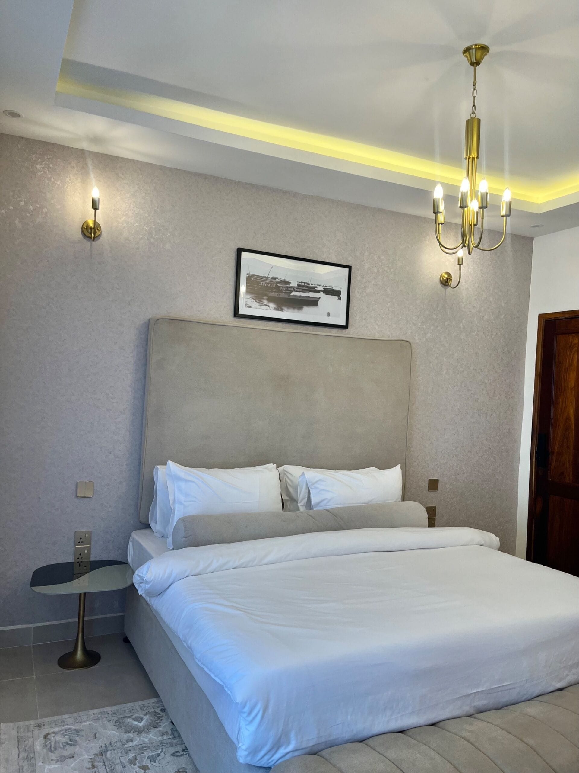 A bedroom with a large bed, a headboard, bedside lamps, a framed picture on the wall, and a chandelier.