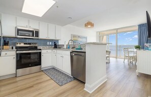A modern kitchen with white cabinets, stainless steel appliances, and a large window overlooking a balcony. - Baywatch1908 Penthouse Oceanfront In/Out pools (North Myrtle Beach)