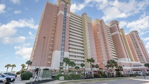 Un hôtel à plusieurs étages au design moderne, doté d’un ascenseur vertical en verre au centre, de nombreux balcons et d’un parking agrémenté de voitures et de palmiers.