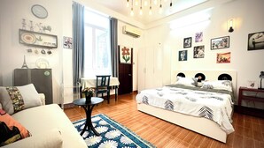Une chambre bien éclairée, avec un lit, un canapé, une petite table et une chaise. Les murs sont décorés et il y a un appareil de climatisation.