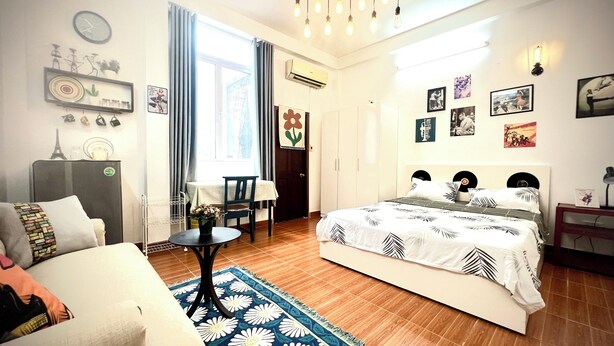 A well-lit bedroom with a bed, a sofa, a small table, and a chair. There are wall decorations and an air conditioning unit. - The Vinh Homestay (Ho Chi Minh City)
