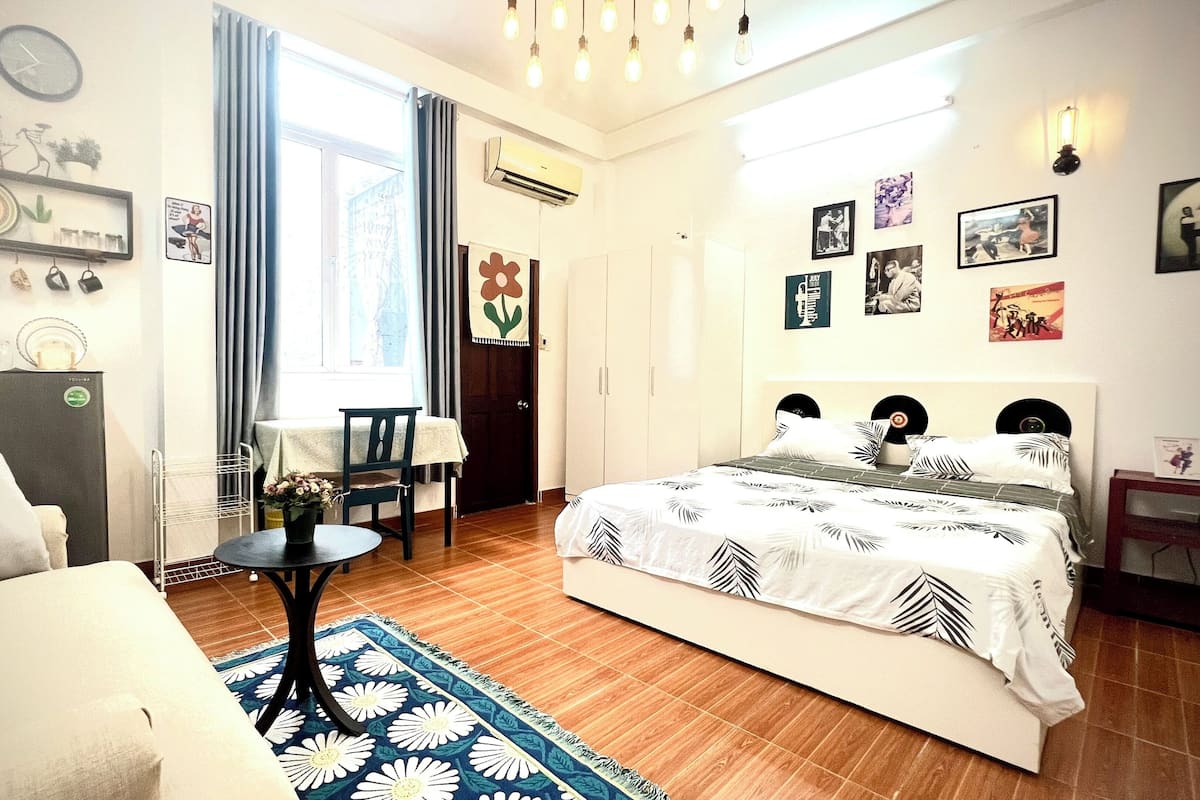 Une chambre bien éclairée, avec un lit, un canapé, une petite table et une chaise. Les murs sont décorés et il y a un appareil de climatisation.