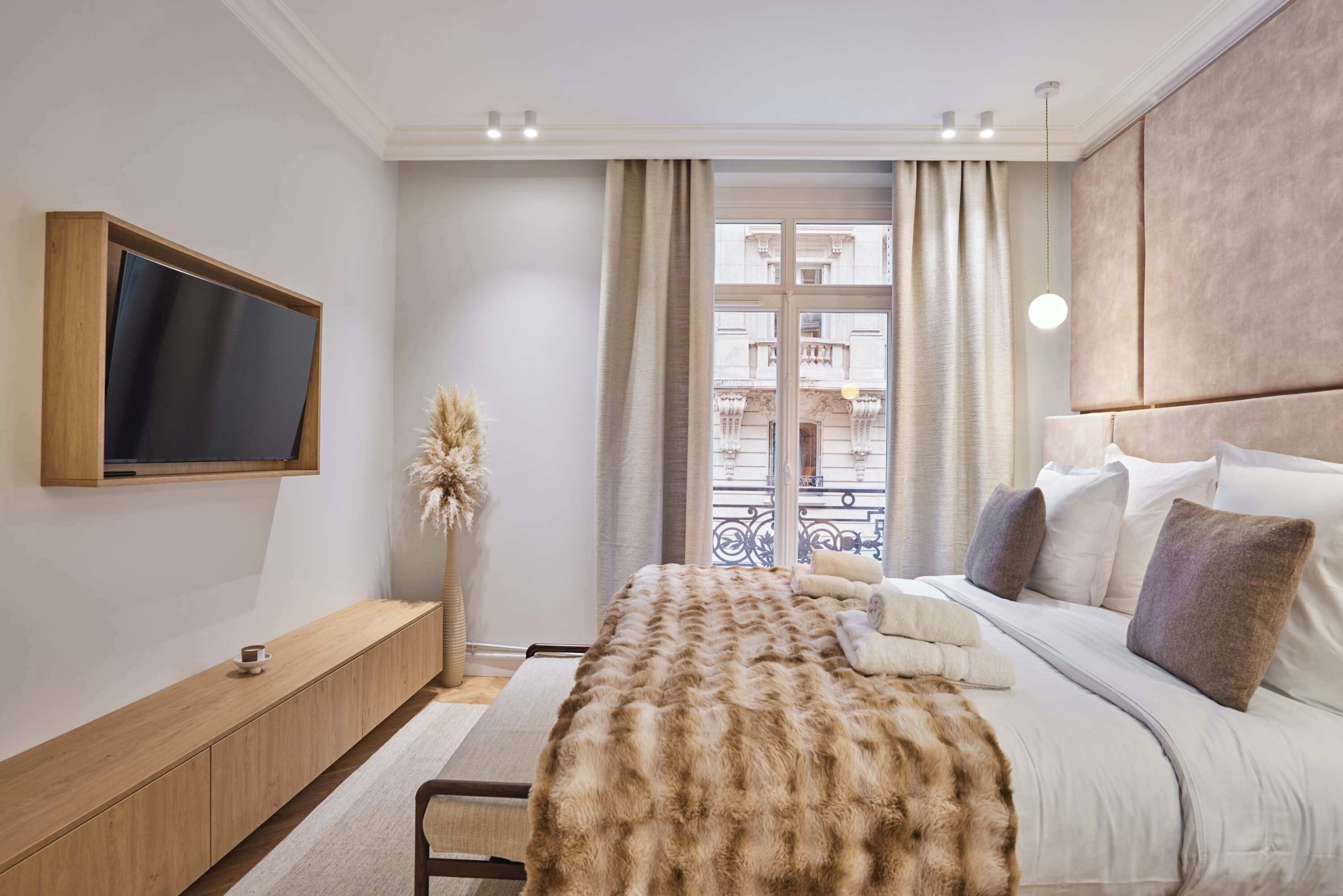 Une chambre moderne avec un grand lit, un téléviseur fixé au mur, une console en bois et une vue sur la ville par la fenêtre.