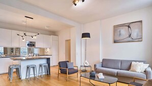 A modern living room with a grey sofa, two armchairs, a coffee table, and a floor lamp. A kitchen with white cabinets and a central island is visible in the background. - Superb apartment Center of Paris Le Marais (Paris)