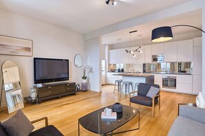 Ein modernes Wohnzimmer mit Flachbildfernseher, Couchtisch und Sofa. Es gibt eine Küchenzeile mit weißen Schränken sowie einen Essbereich mit Barhocker.