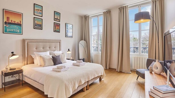 A well-lit bedroom with a large bed, a bedside table with a lamp, a chair, and a desk with books and a candle. - Superb apartment Center of Paris Le Marais (Paris)