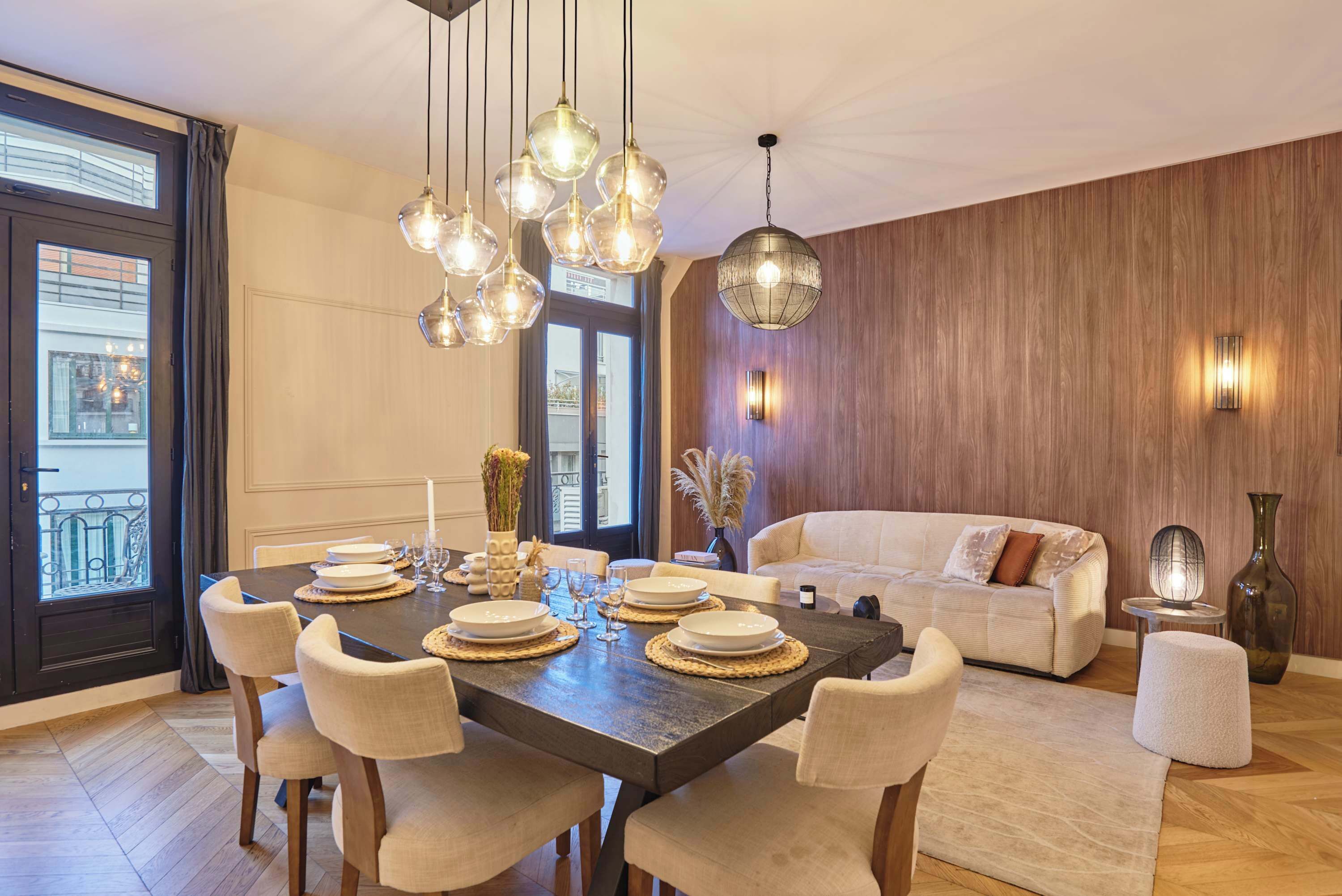 A dining area with a large table set for six, surrounded by chairs. There is a sofa and a decorative vase in the background.