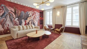A living room with a large abstract mural, a beige sofa, two round coffee tables, and a red carpet. - Luxurious AC Apartment Louvre Museum Saint Honoré (Paris)