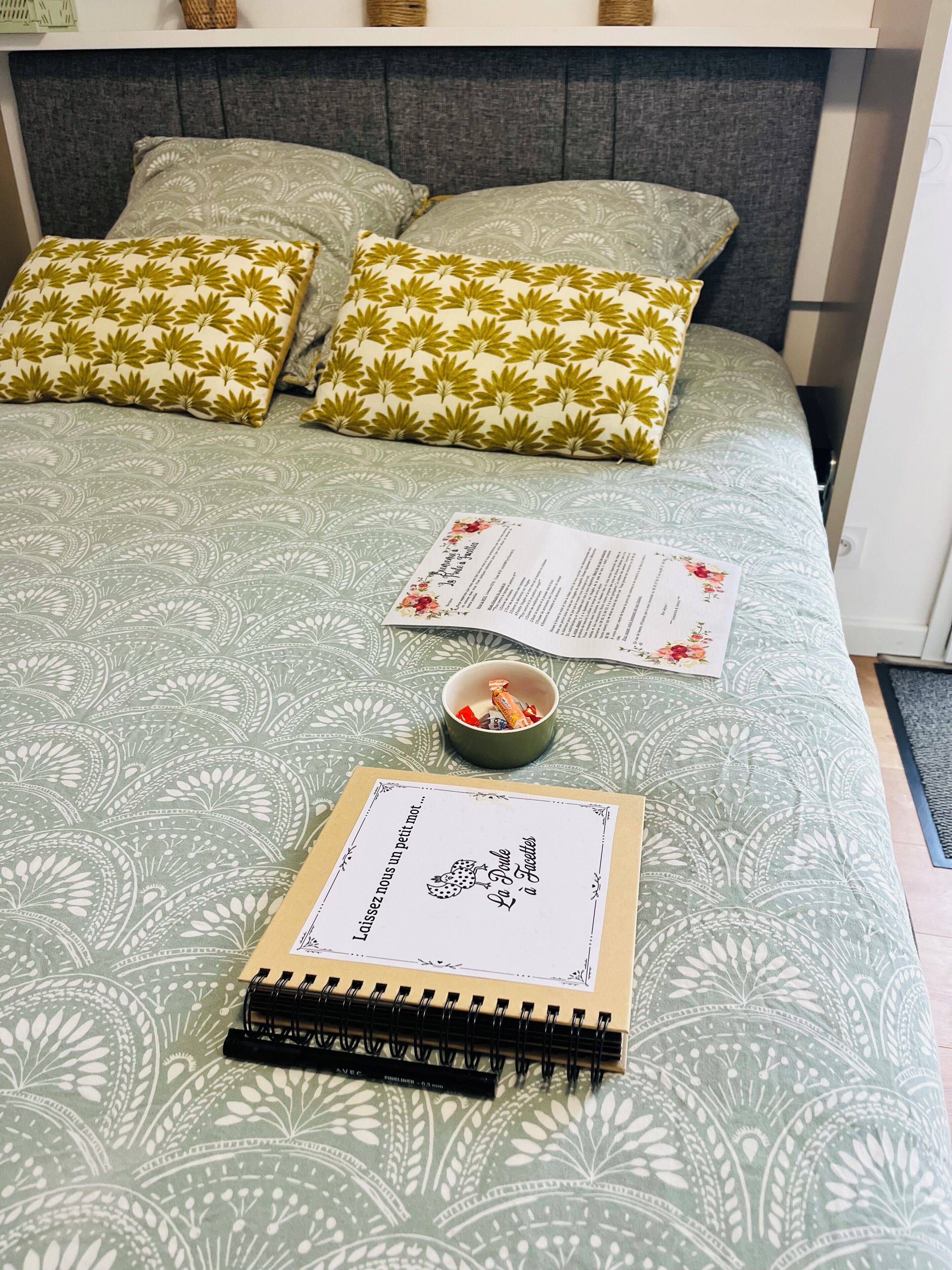 Ein ordentlich gemachtes Bett mit gemusterten Betttextilien, eine kleine Tasse mit einer Leckerei und ein Notizbuch auf dem Bett.
