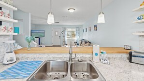 A modern kitchen with a double sink, a blender, and a toaster. There are hanging lights, a wooden countertop, and a view of the living room in the background. - Stay at The Salty Palm! 2BR 1.5ba Sleeps 7 (Panama City Beach)