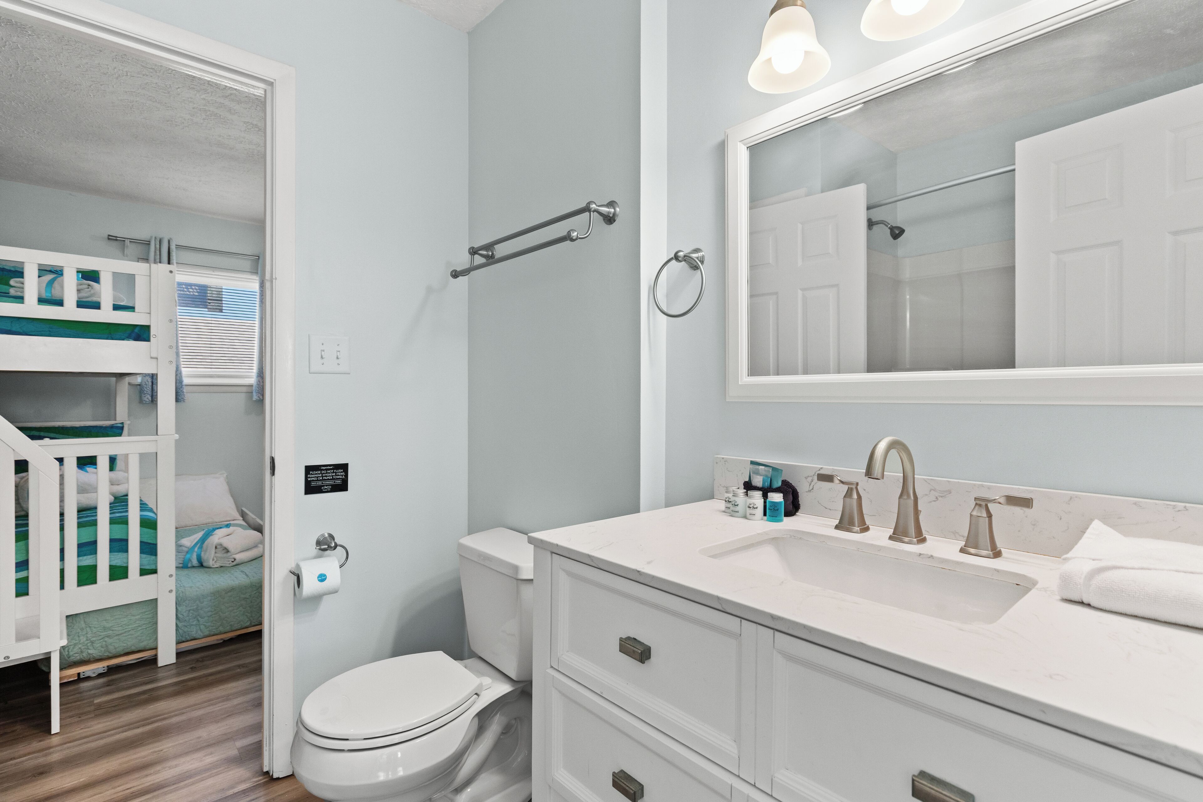 A bathroom with a white sink, a mirror, and a toilet. There is a bunk bed in the adjacent room.