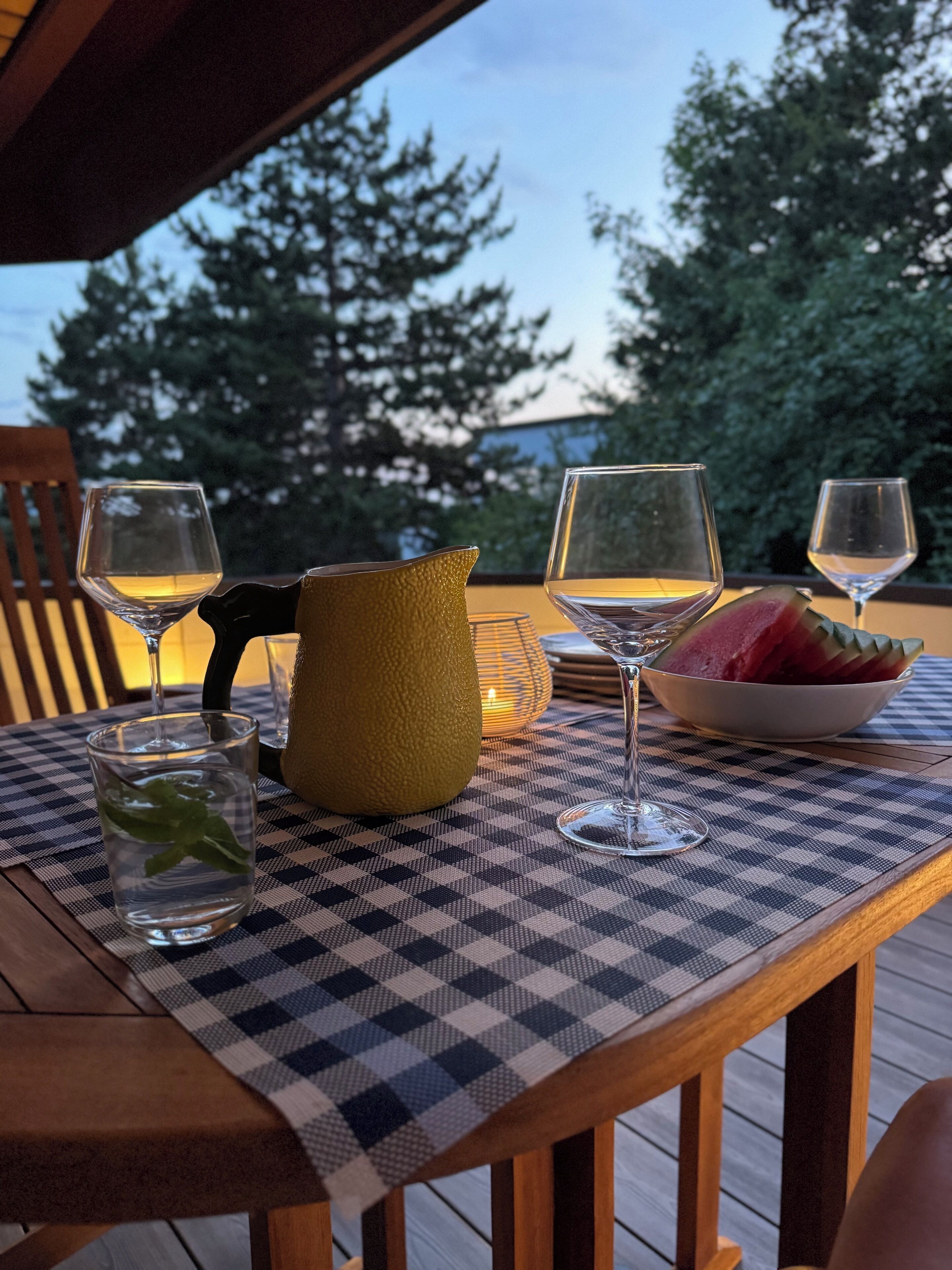 A table set with wine glasses, a pitcher, and a bowl of fruit on a checkered tablecloth.
