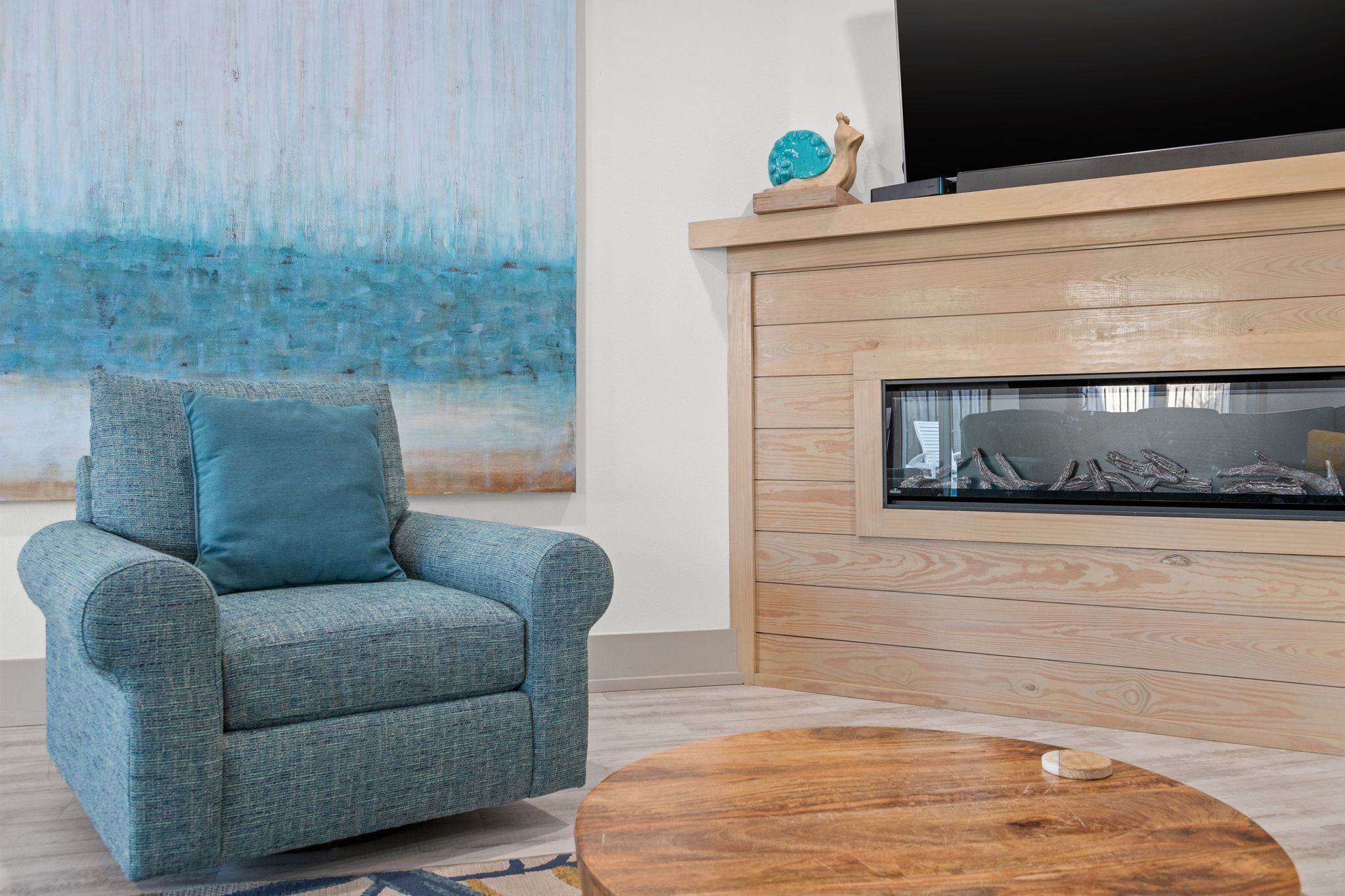 A modern living room with a blue armchair, a wooden coffee table, a fireplace, and a large abstract painting.