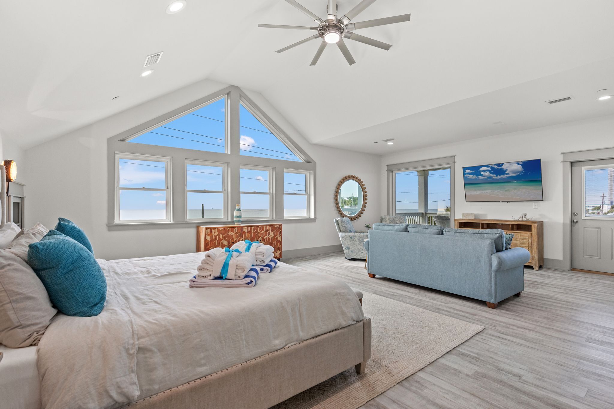 A spacious bedroom with a large bed, a ceiling fan, and a view of the sky through large windows.
