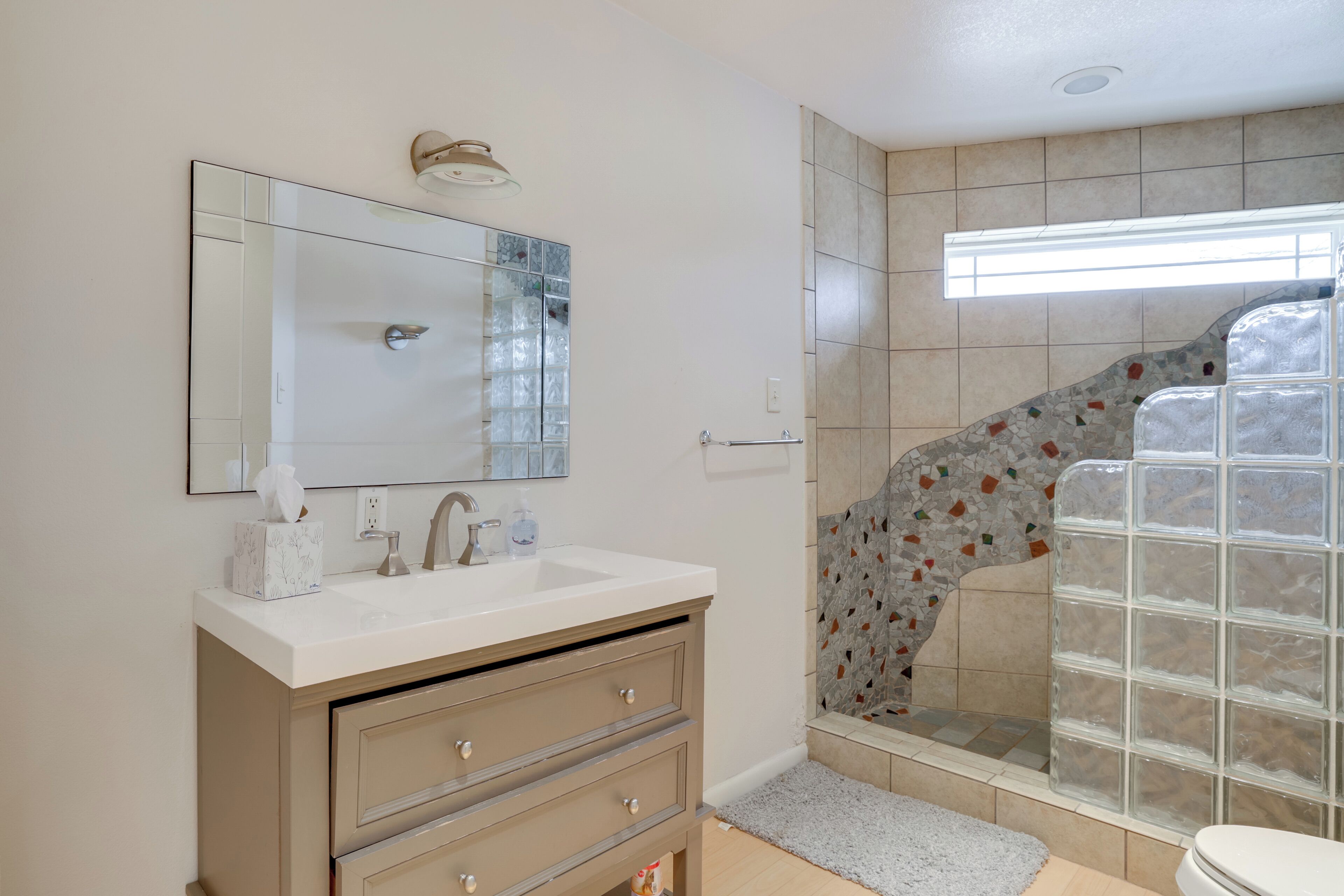 Une salle de bain avec un lavabo blanc, un meuble-lavabo beige et une cabine de douche en verre ornée de mosaïques.