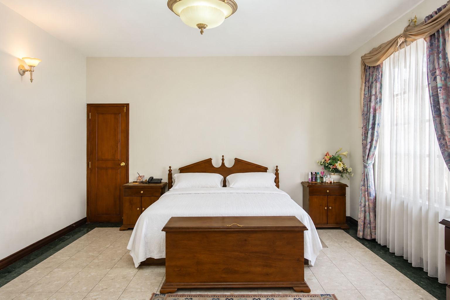 A bedroom with a wooden bed, bedside tables, a nightstand, a wooden bench, a door, and a window with curtains.