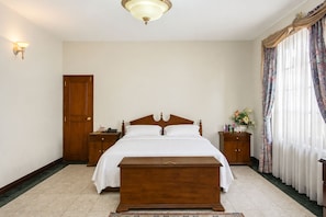 A bedroom with a wooden bed, bedside tables, a nightstand, a wooden bench, a door, and a window with curtains.