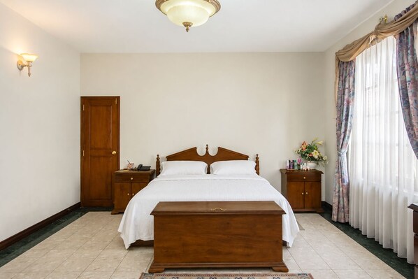 A bedroom with a wooden bed, bedside tables, a nightstand, a wooden bench, a door, and a window with curtains.