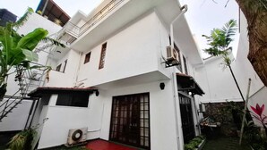 A white two-story house with a red-tiled roof, a large sliding glass door, and a small garden area with plants and a tree.