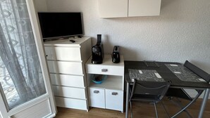 Quarto pequeno e funcional com guarda-roupa branco, escrivaninha com cadeira, televisão e torradeira.