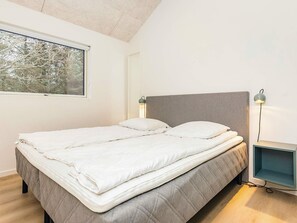 A neatly made bed with a grey headboard and white bedding, a bedside table with a lamp, and a window with a view of trees. - 5 Star Holiday Home in Blokhus (Blokhus)
