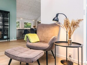 A modern living room with a tufted ottoman, a chair with a throw blanket, a side table with a lamp, and a vase with dried plants. - 5 Star Holiday Home in Blokhus (Blokhus)