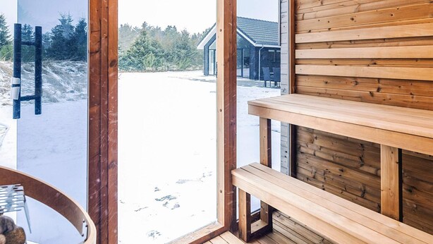 Een houten interieur met een bank en een tafel, een glazen deur die naar buiten leidt, en uitzicht op een gebouw en bomen.