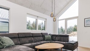 Een moderne woonkamer met een hoekbank, een ronde salontafel en grote ramen met uitzicht op bomen en een besneeerd landschap.