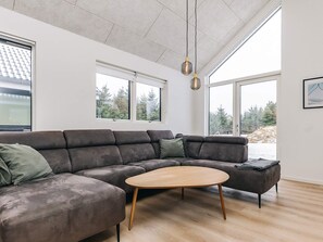 Ein modernes Wohnzimmer mit einer Eckcouch, einem runden Couchtisch und großen Fenstern mit Blick auf Bäume und eine verschneite Landschaft.