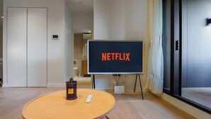 Huoneessa on pyöreä puupöytä, televisiossa näkyy teksti ”NETFLIX”, ja pöydällä on pieni valkoinen laite.
