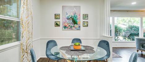 A dining area with a glass table surrounded by velvet chairs, a central plant, and a peacock painting on the wall.