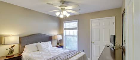 Une chambre à coucher comprenant un lit, des tables de chevet, un ventilateur de plafond, une fenêtre et un téléviseur.