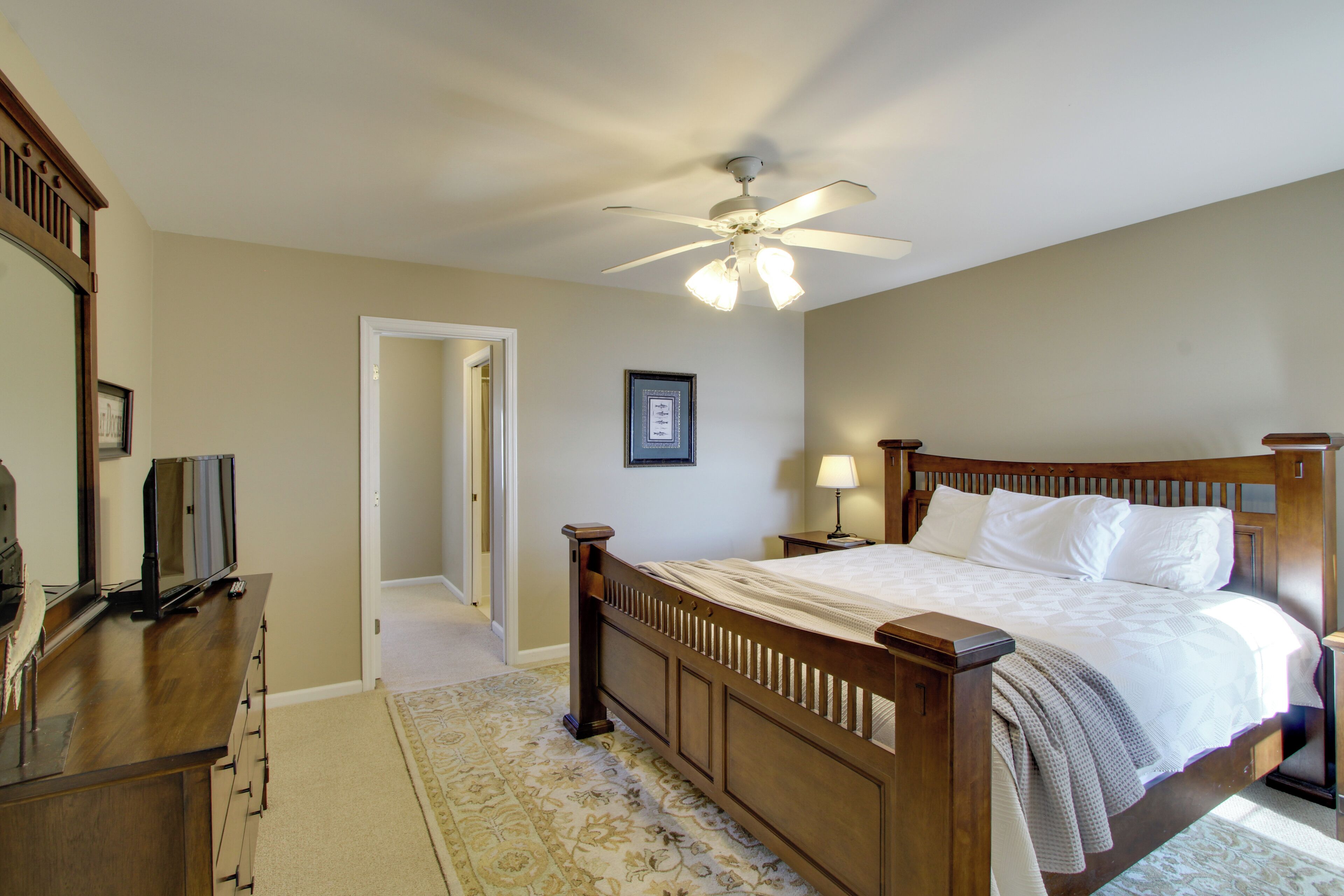 A bedroom with a wooden bed, a ceiling fan, a television on a dresser, and a framed picture on the wall.
