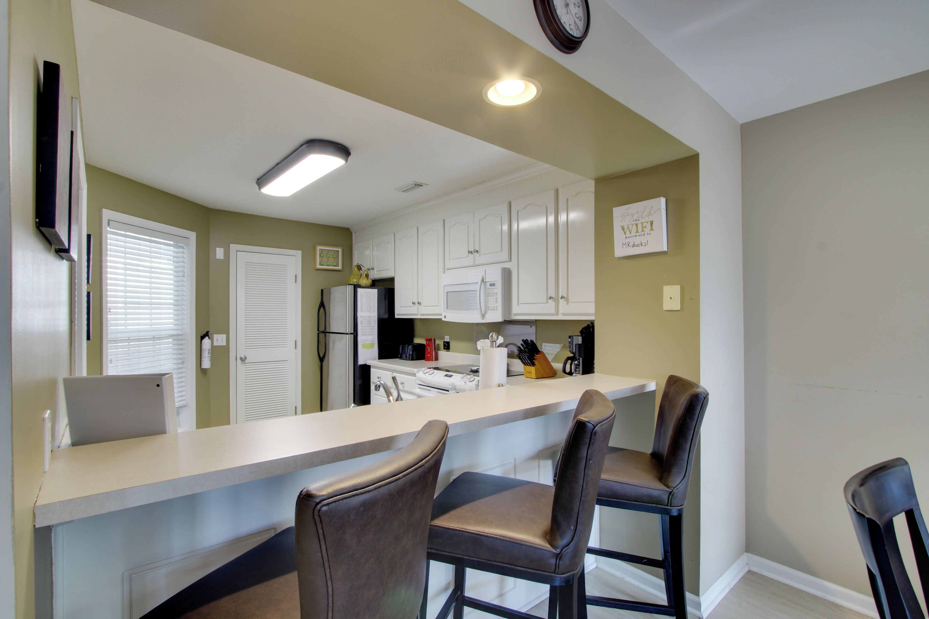 A kitchen with white cabinets, a microwave, and a sink. There are two bar stools and a clock on the wall.