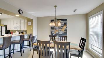 A dining area with a wooden table and chairs, a kitchen in the background, and a wall-mounted clock.