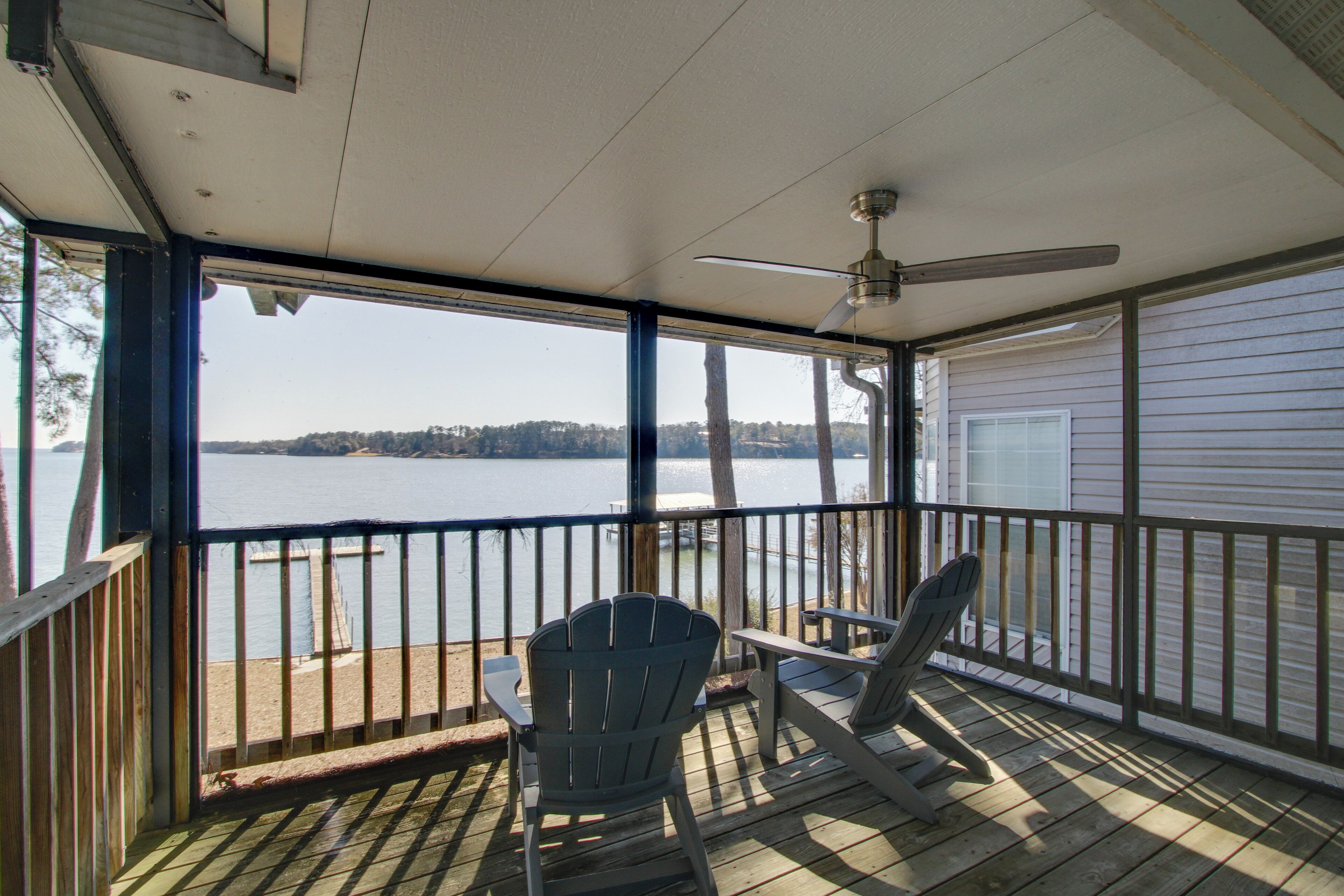 A covered deck with two Adirondack chairs and a table, overlooking a lake with a distant shoreline.