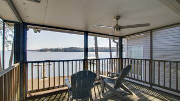 A covered deck with two Adirondack chairs and a table, overlooking a lake with a distant shoreline.
