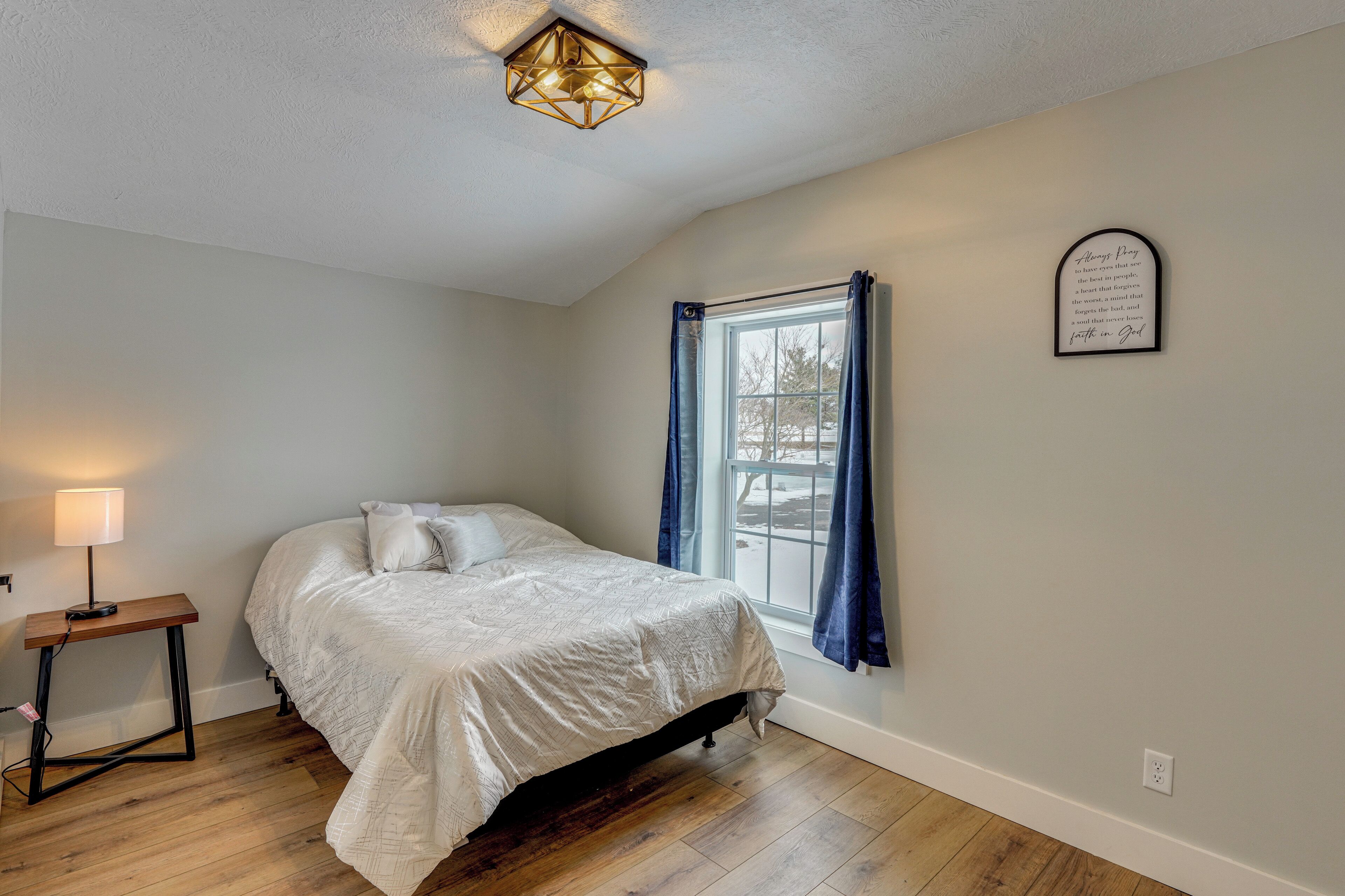 A bedroom with a bed, a bedside table, a lamp, a window with curtains, and a wall-mounted light fixture.