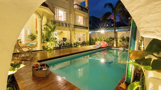 An indoor swimming pool with a wooden deck, surrounded by plants and a building with balconies. - AnNam Village 4 (Vũng Tàu)