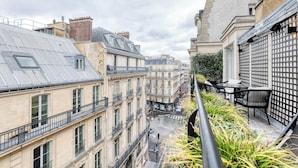 Una estrecha calle urbana flanqueada por edificios con arquitectura clásica europea, que incluyen balcones y un jardín en la azotea.